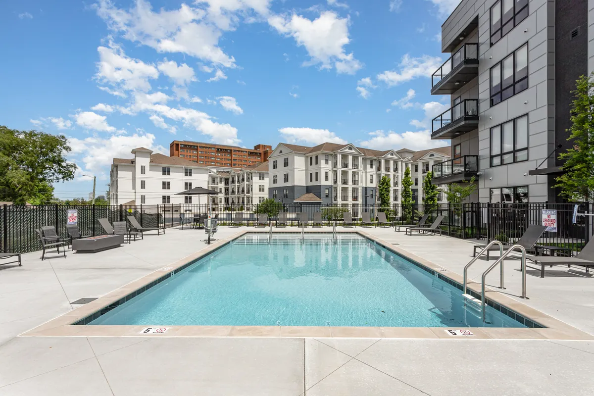 This image presents a sparkling swimming pool surrounded by lounge chairs and modern apartment buildings, all set amidst lush greenery. The inviting water and comfortable seating convey a sense of leisure and relaxation, while the architectural backdrop subtly hints at the upscale, resort-style ambiance.