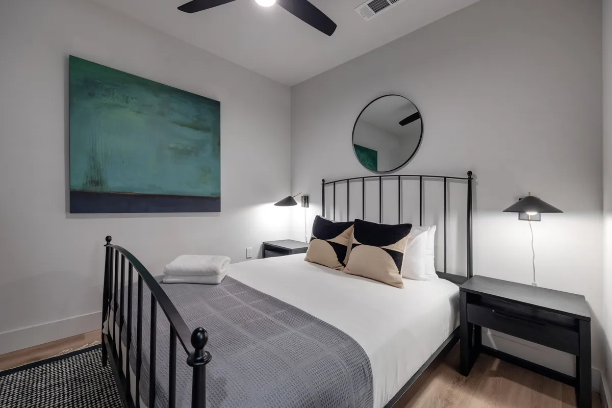 A serene bedroom with a comfortable bed, round mirror above the headboard, abstract wall art, nightstands on each side, and soft table lamps, all in a tranquil, inviting palette.