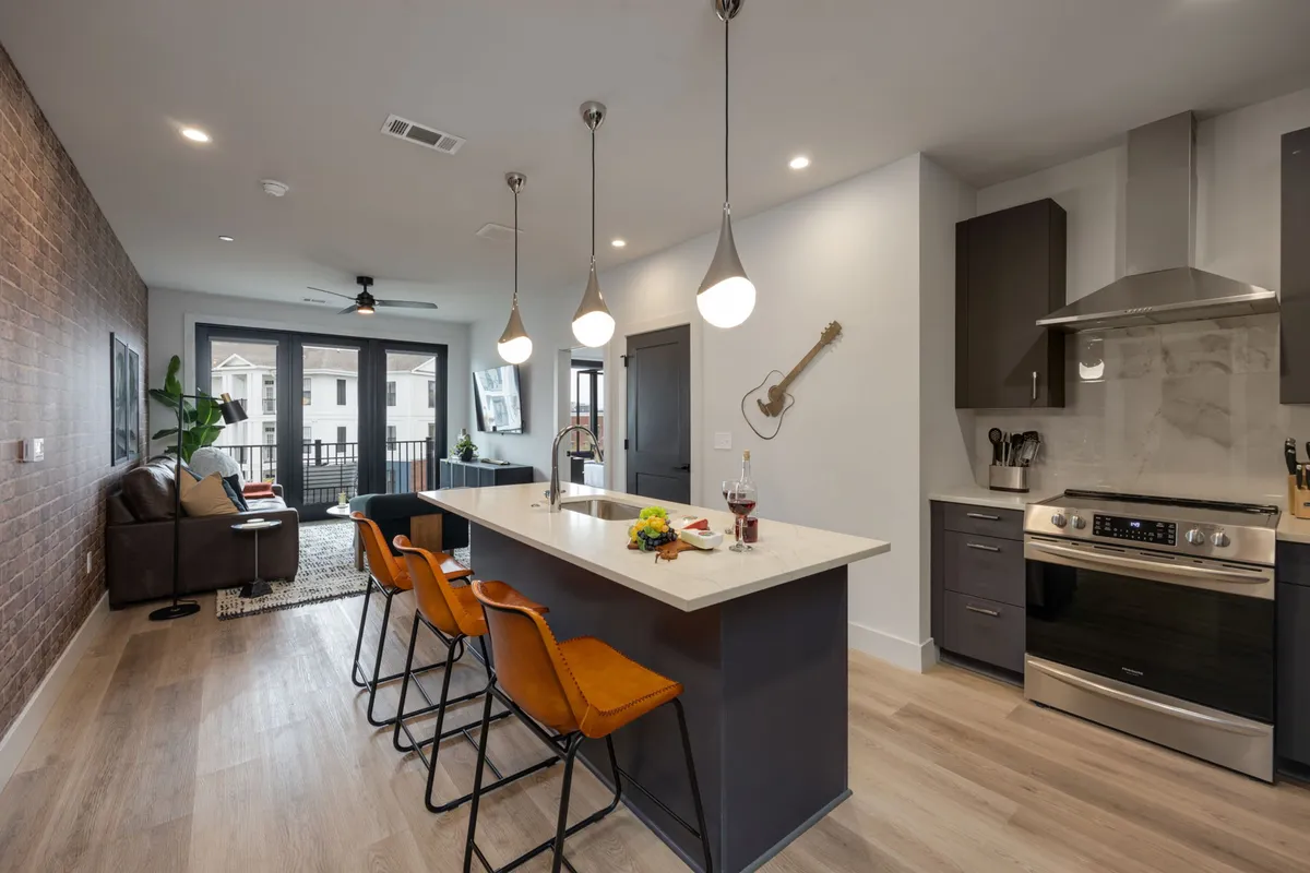 An open-concept living space seamlessly joining the kitchen and living room. The kitchen features a spacious island with bar stools, sleek cabinetry, and a guitar on the wall, while the adjacent area has a comfortable sofa and large windows letting in abundant sunlight. The overall atmosphere is contemporary and welcoming, perfect for gatherings and relaxation.