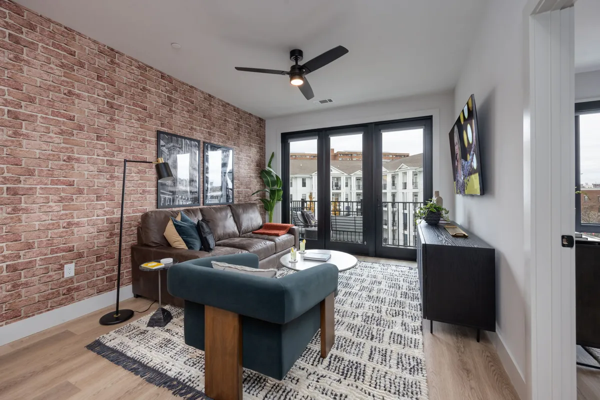 The image showcases a stylish, contemporary living room featuring a striking brick accent wall, a plush leather sofa, and expansive glass doors that open to a private balcony. The space is bright and inviting, perfect for unwinding, hosting friends, or enjoying a quiet evening. The blend of modern design and cozy touches sets an inviting mood for guests.