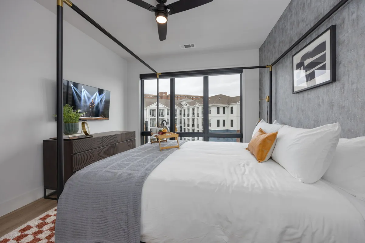 A stylish and spacious bedroom featuring a cozy canopy king bed, a TV mounted on the wall, large windows flooding the space with natural light, and a decorative tray set atop crisp bed linens.