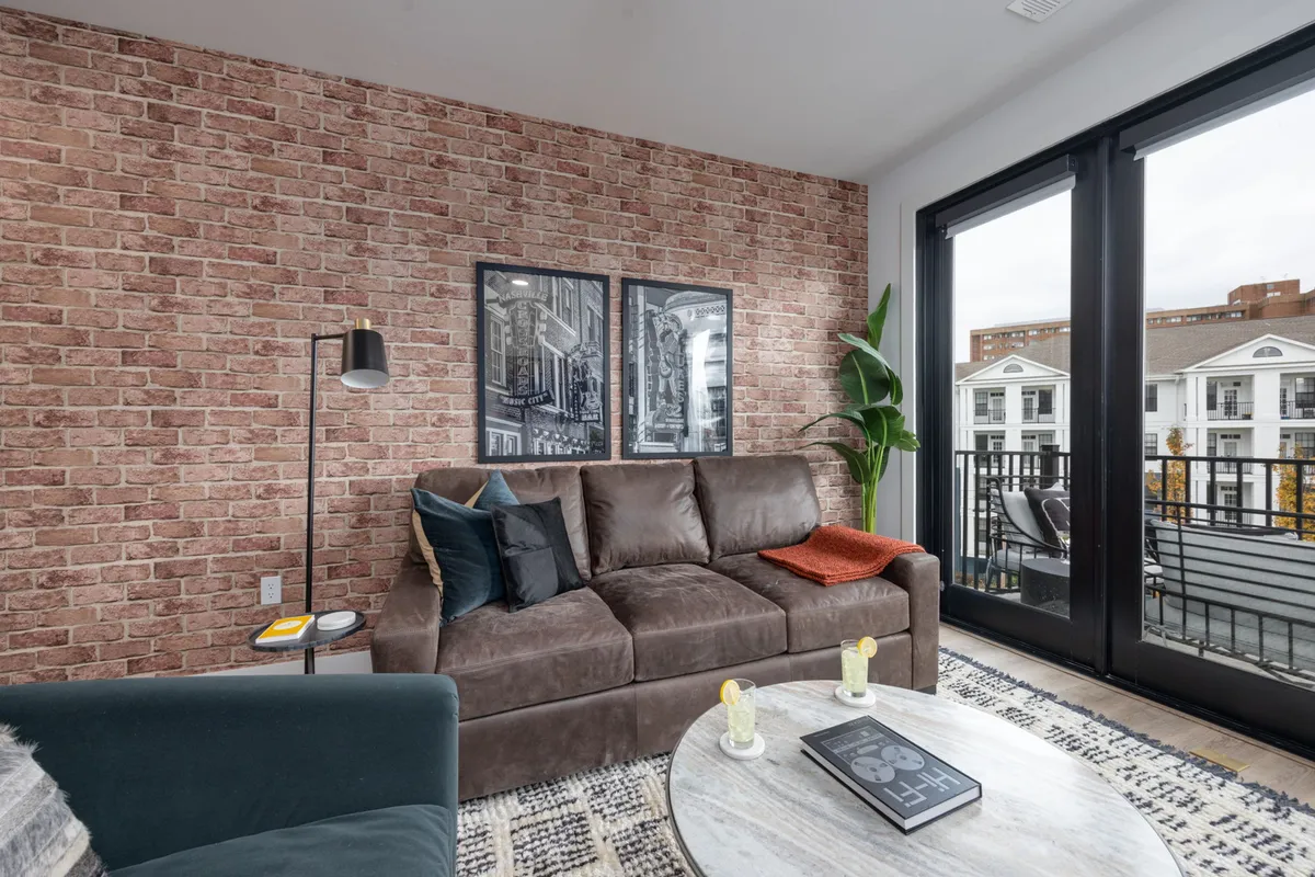 A stylish living area with a brown sofa, brick wall, and a coffee table, with a clear view out to the balcony. The inviting mood suggests seamless indoor-outdoor living, and you can see the balcony as an extension of the relaxation space.